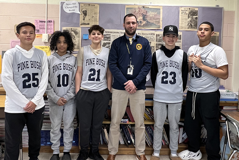 A man in the center with a blue jacket that has a gold PB logo. He has short dark hair and a close beard and mustache. There are three boys on his left and two on his right,, all in basketball jerseys.