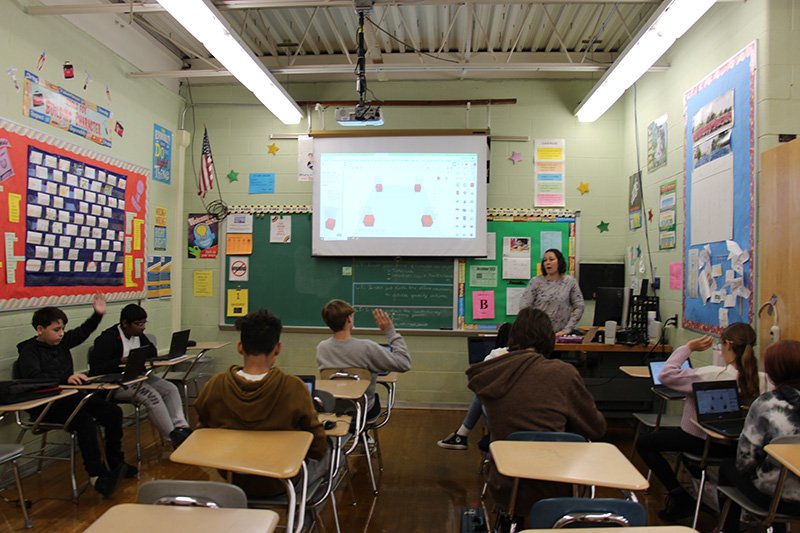 A classroom from the back with a large screen overhead. Students are sitting at desks and a woman is at the front talking.