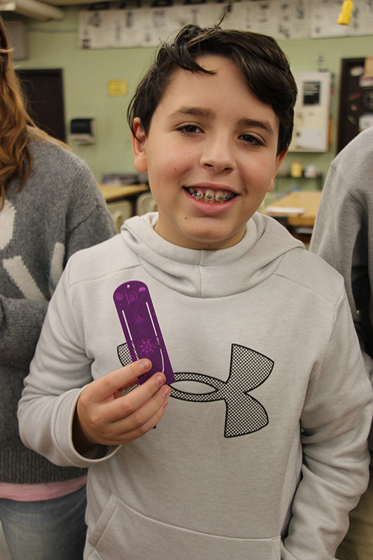 A middle-school boy with short dark hair smiles. He is wearing a gray sweatshirt and holding a purple bookmark he made.
