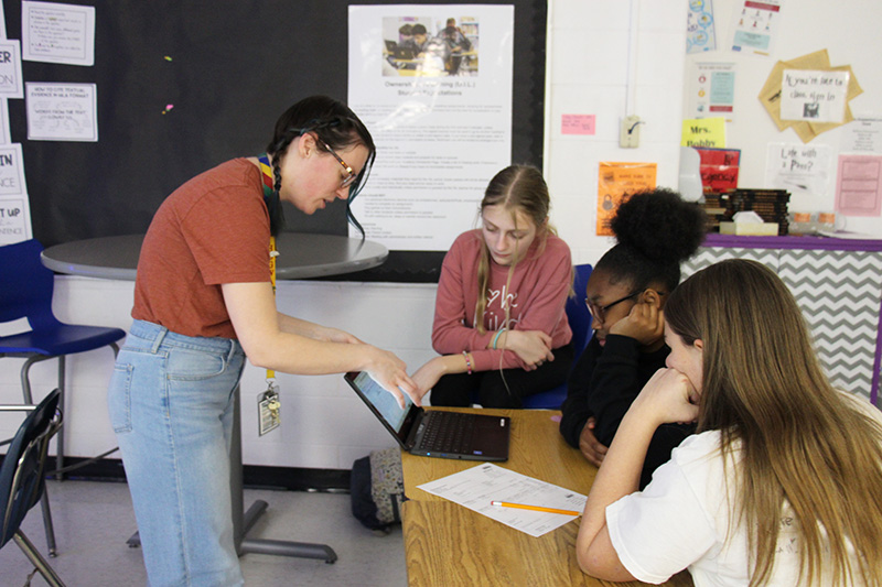 A woman leans in and talks to three middle school girls who are working on a Chromebook.