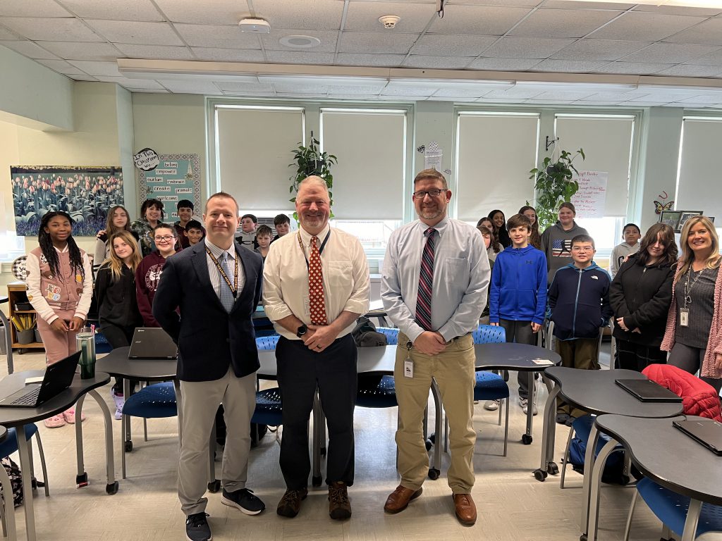 Three men stand in front, each wearing button-down shirts and ties. They are smiling. Behind them are middle school students.