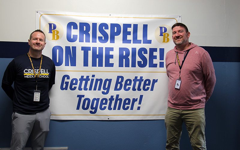 Two men smile and stand on either side of a sign that says Crispell On The Rise! Getting Better Together!