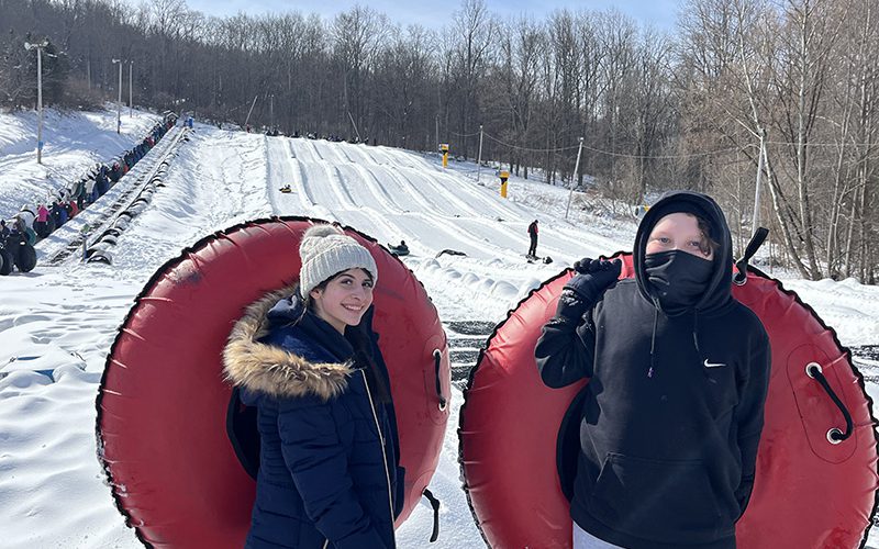 Two middle school students hold big red snow tubes with a mountain behind them.
