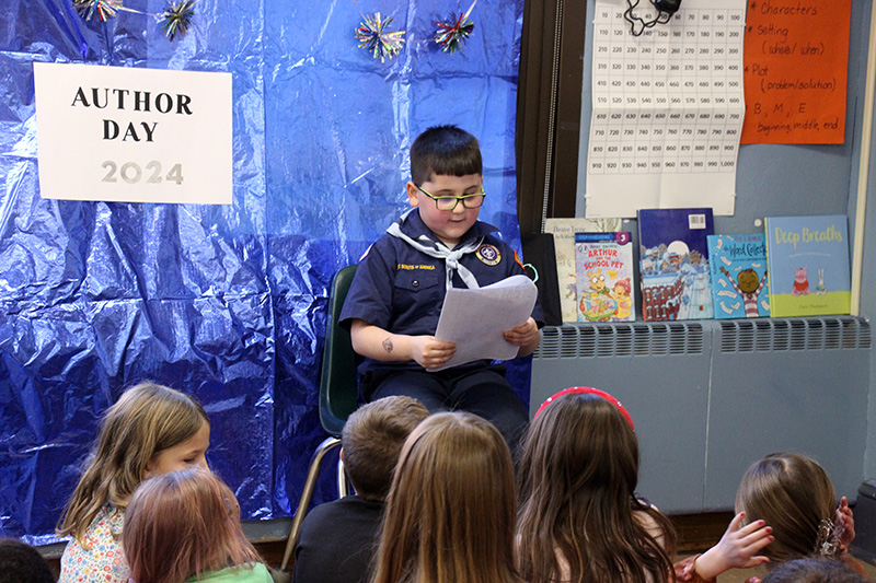 A third-grade boy, dressed in his navy blue cub scout uniform, reads his writing to a group of students.