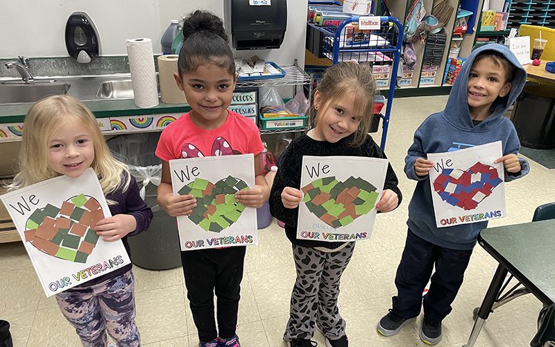 Four kindergarten students stand next to each other smiling. They are each holding a valentine they made That says We Heart our veterans. The hearts are filled in with cut up pieces of paper is green and brown or red, white and blue.