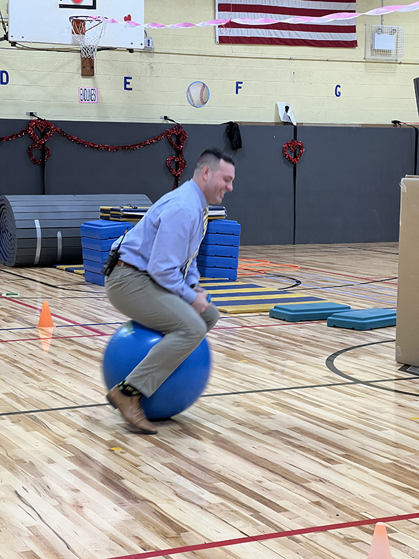 A man in a blue dress shirt and khaki pants sits on a blue hippity hop ball and bounces.