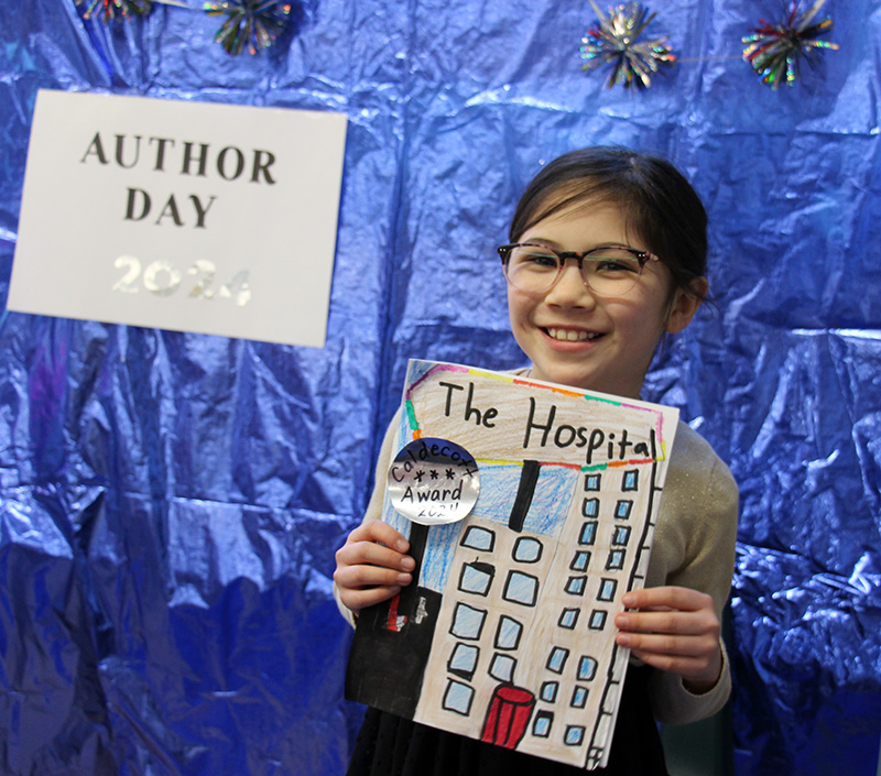 A girl with long dark hair pulled  back into a ponytail and wearing glasses, smiles as she holds up her book called The Hospital. She is smiling. The background is shiny blue with a sign that says Author Day.