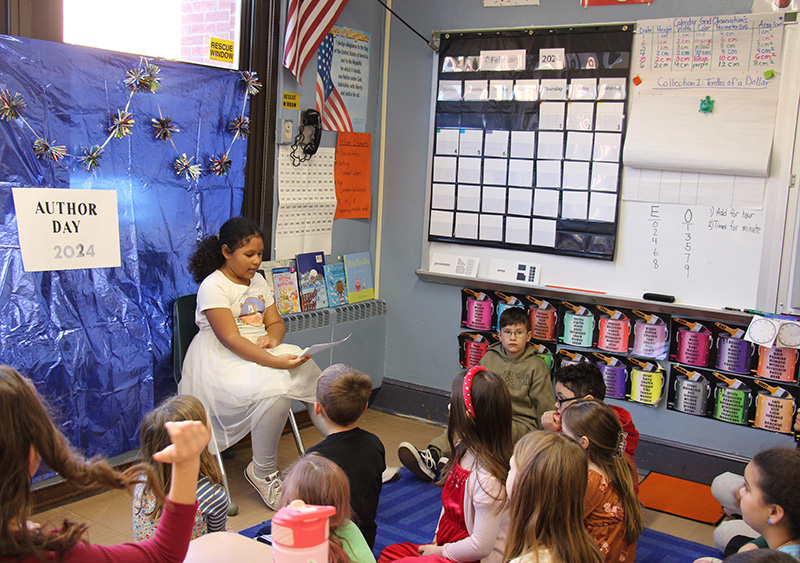 A girl with long dark hair dressed in a fancy white dress, reads from her book to her classmates sitting on the floor in front of her.