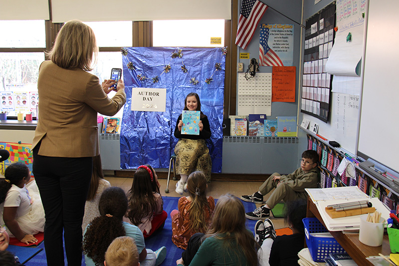 A girl sits in front of a blue wall holdin gup her book. A group of her classmates are sitting on the floor in front of her and a woman is taking a picture of her.