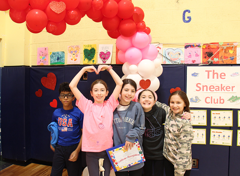 A group of five elementary students stand in front of a pine and red balloon heart smiling.
