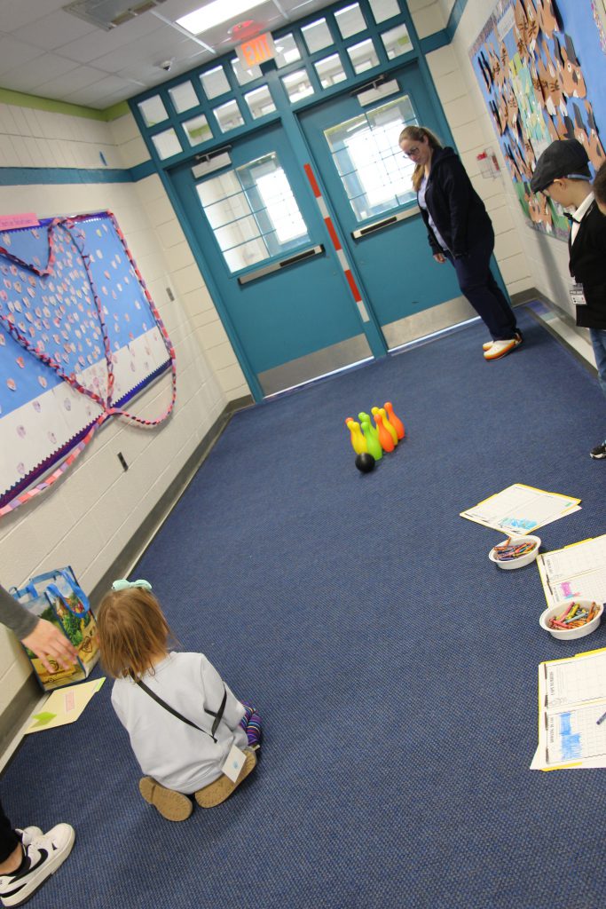 A kindergarten student stands watching a ball roll through the hallway to 10 plastic bowling pins down a blue carpet.