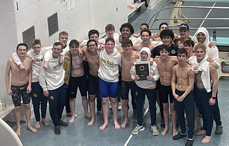 A group of 22 high school boys  stand by a pool, many in bathing suits. They are smiling. A boy in the front holds a plaque.