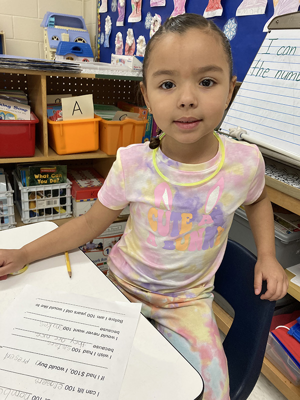 A kindergarten girl with a purple and pink shirt on smiles as she works on a worksheet.
