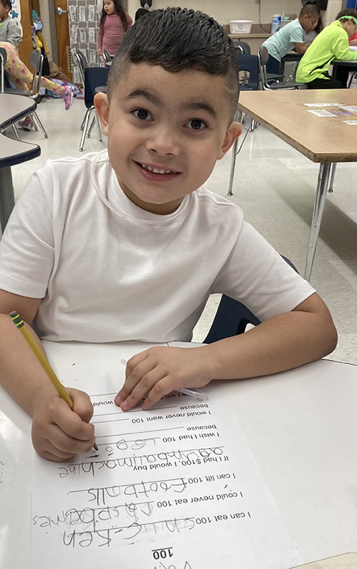 a kindergarten boy in a white tshirt with short dark hair smiles as he works on a worksheet.