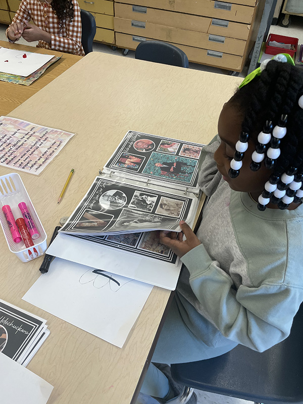 A girl with white beads in her hair looks through binders that have artwork in them.