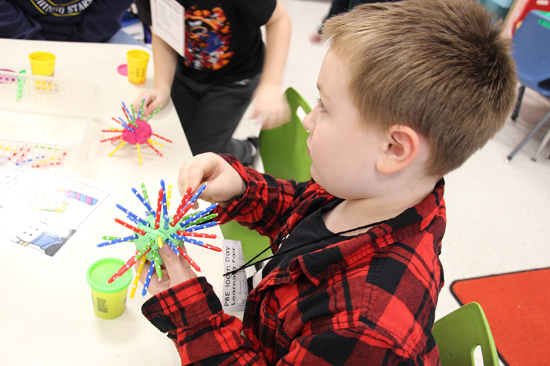 A boy with short light-colored hair, wearing a red and black plaid shirt, puts candles into a ball of Playdoh.