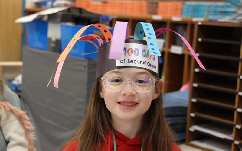 A second grade girl with long brown hair and glasses wears a hat made of paper that says 100 days of second grade. There are strips of paper with dots on them flopping over the hat.,