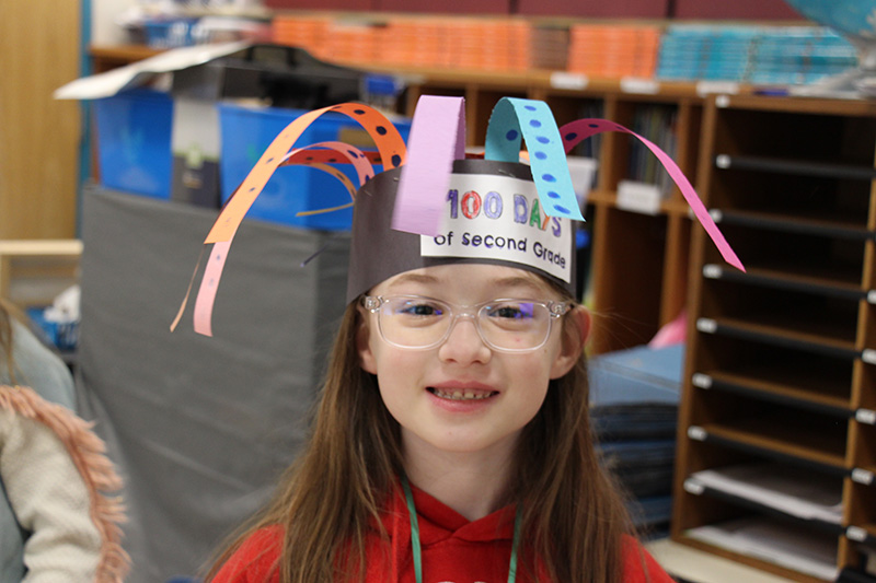 A second-grade girl with long brown hair and glasses smiles with a paper hat on her head that says 100 days of second grade.