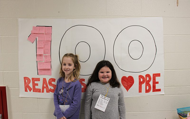 Two elementary girls smile as they stand in front of a big poster that says 100 Reasons to love PBE