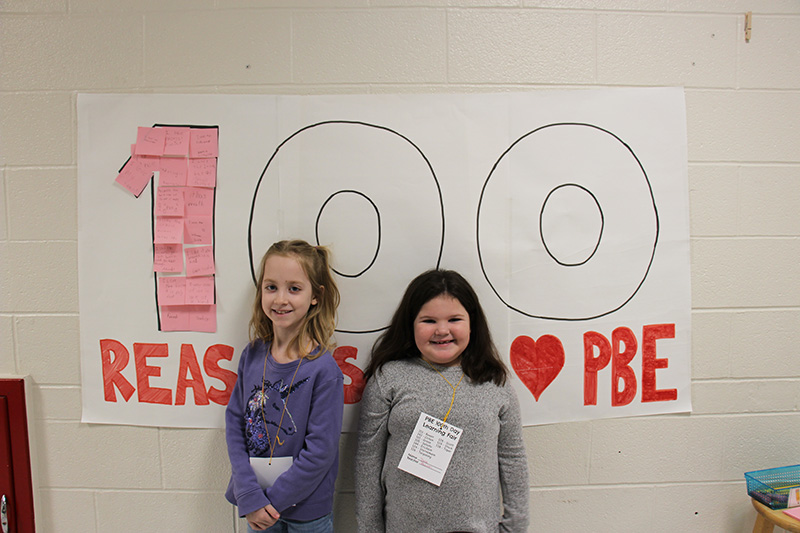 Two elementary age girls stand infront of a big poster that says 100 reasons to love PBE