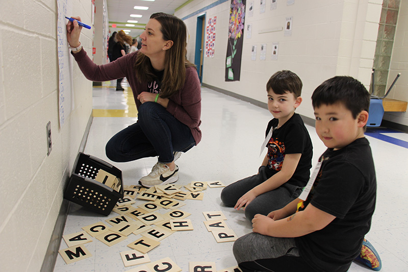 Two elementary boys sit with squares that have letters on them. They are creating words from the squares and a woman bends in front of them writing the words on a large piece of paper.