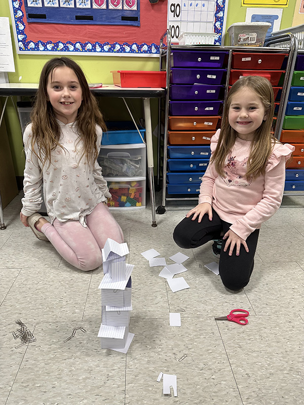 Two elementary age girls sit on the floor building a tower made of index cards and paper clips. They both have long brown hair and are smiling.