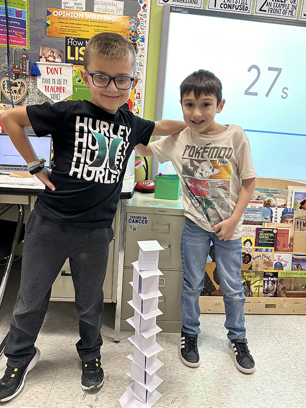 Two elementary age boys stand proudly over a tower they built out of index cards and paper clips. They have one arm around each other and the other on their hips.