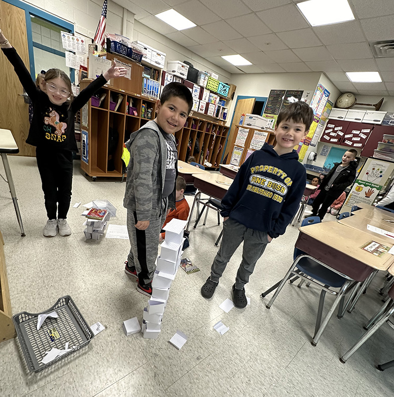 Three elementary students stand looking at a tower they built of index cards and paper clips. The girl on the left has her arms up high with a jubilant smile on her face. The two boys are smiling.