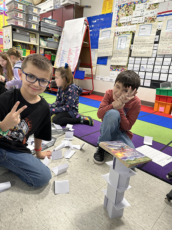 Two elementary boys sit on the floor near a tower they just built from index cards and paper clips. There is a book on top. The boy on the left is giving a peace sign.