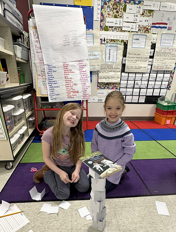 Two elementary age girls sit on the floor behind a tower they made of index cards and paper clips. They both have long brown hair and are smiling.