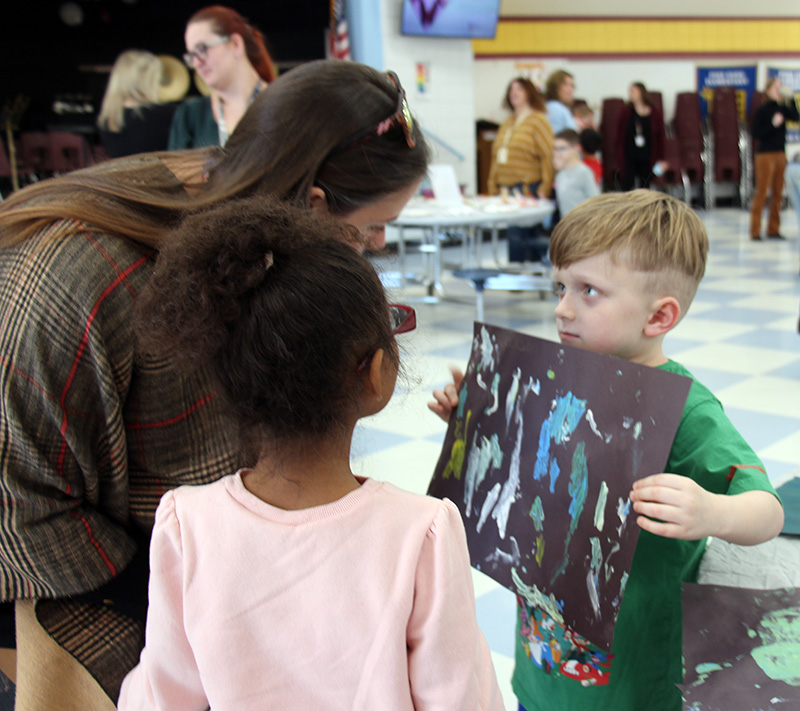 A young elementary boy with short hair holds up his painting for two people to look at.