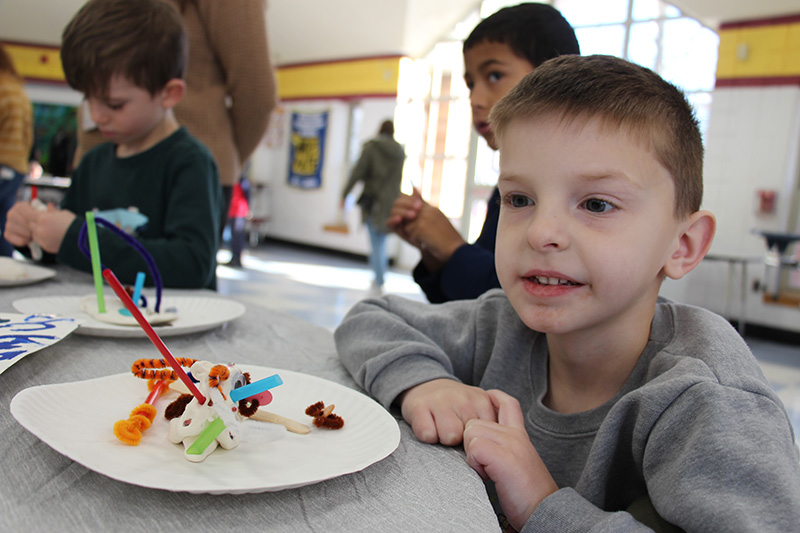 An elementary boy with short light hair sits next to a piece f 3D art he created.