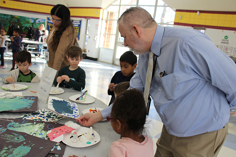 A man in a blue shirt and tie leans over to see artwork by a young elementary student.
