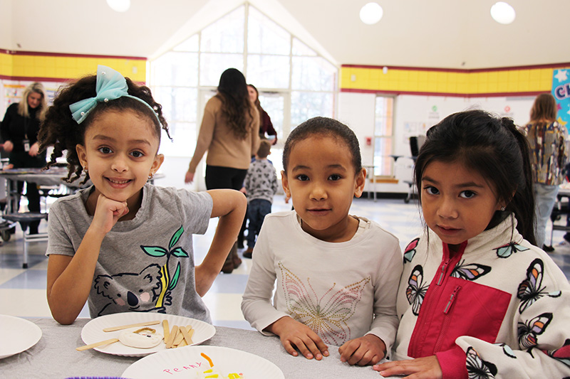 Three young elementary students stand together near a table of artwork.