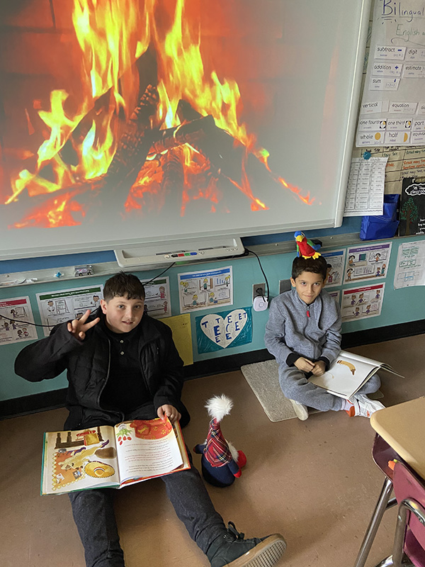 Two boys sit on a floor reading books with a large photo of a fireplace behind them.