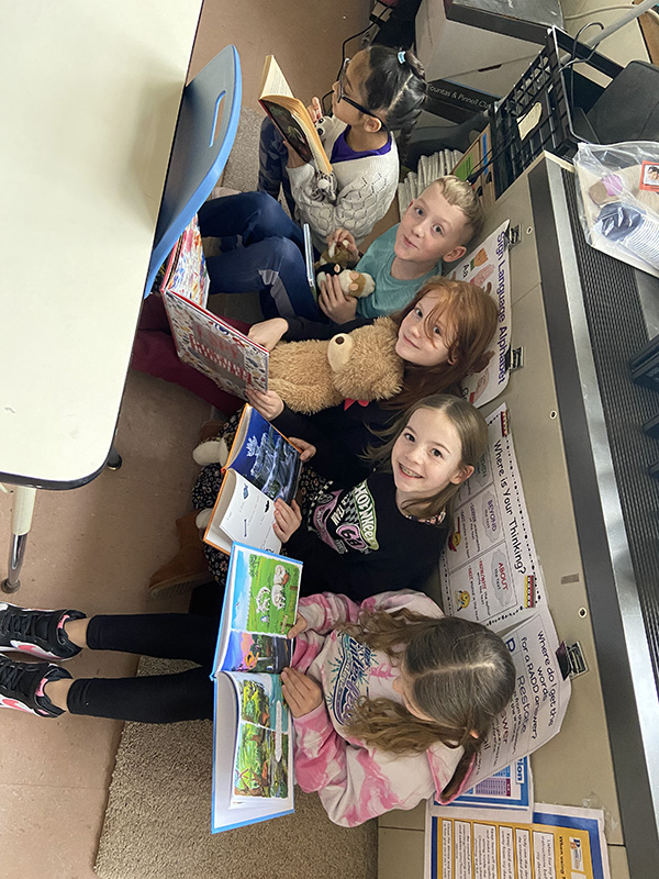 Five fourth-grade girls sit on the floor in a row reading their books and looking up toward the camera.