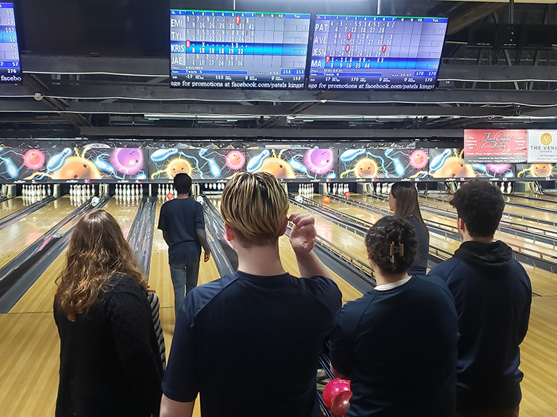 A group of high school kids stand with their backs to the camera watching a teammate bowl.