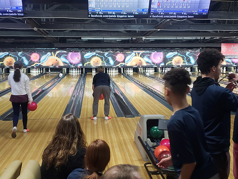 The setting is a bowling alley. There is a person bending to bowl with others watching.