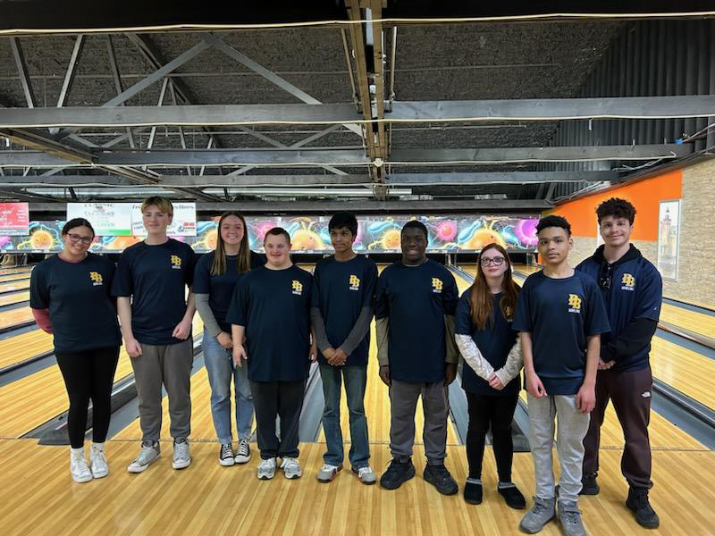 A group of nine high school kids stand together with a bowling lane in the back.