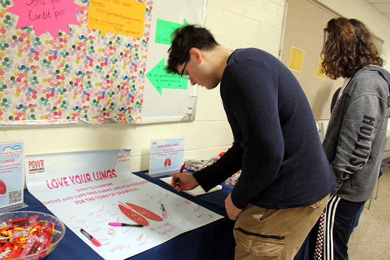A high school student signs his name to a red and white poster.