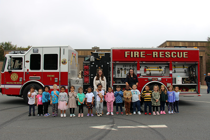 Two women stand in front of a large red and white fire truck with a group of little elementary students in front.