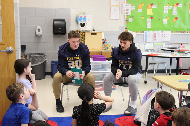 Two football players read a book to younger elementary students who are sitting on a rub.