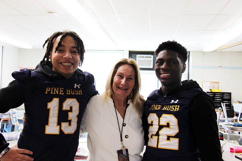 Two high school football players wearing their jerseys stand with their arms around a woman who used to be their teacher. They are all smiling broadly.