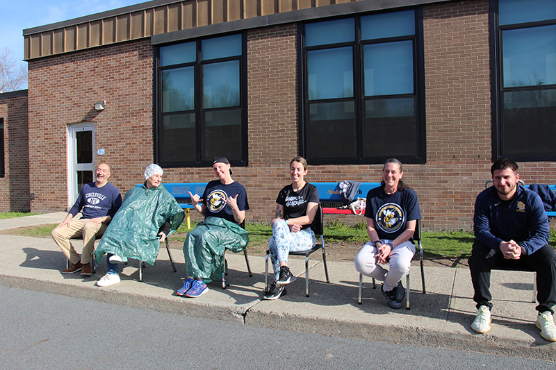 Six adults sit in chairs outside a school building.