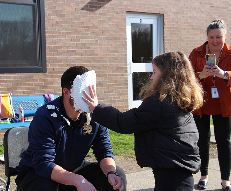 A girl with long dark hair smushes a plate of whipped cream into the face of a man sitting down.