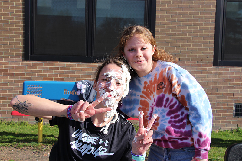 A woman with whipped cream all over her face gives peace signs as a girl with a tie dye tshirt leans in and smiles.