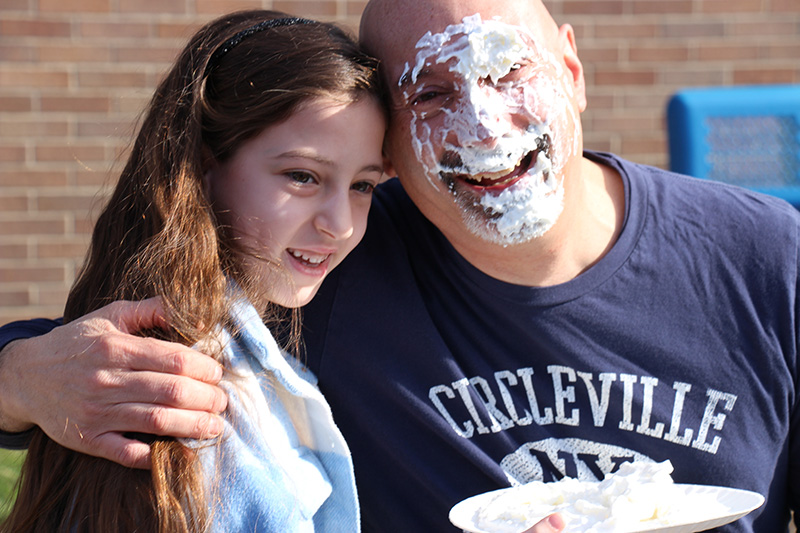 A man wearing a blue shirt has whipped cream all over his face. He is smiling and has his arm around a girl with long dark hair.