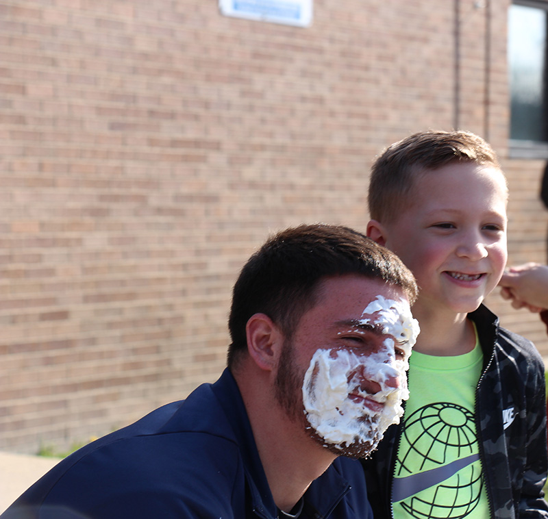 A man with whipped cream all over his face smiles with a young boy who put it there.