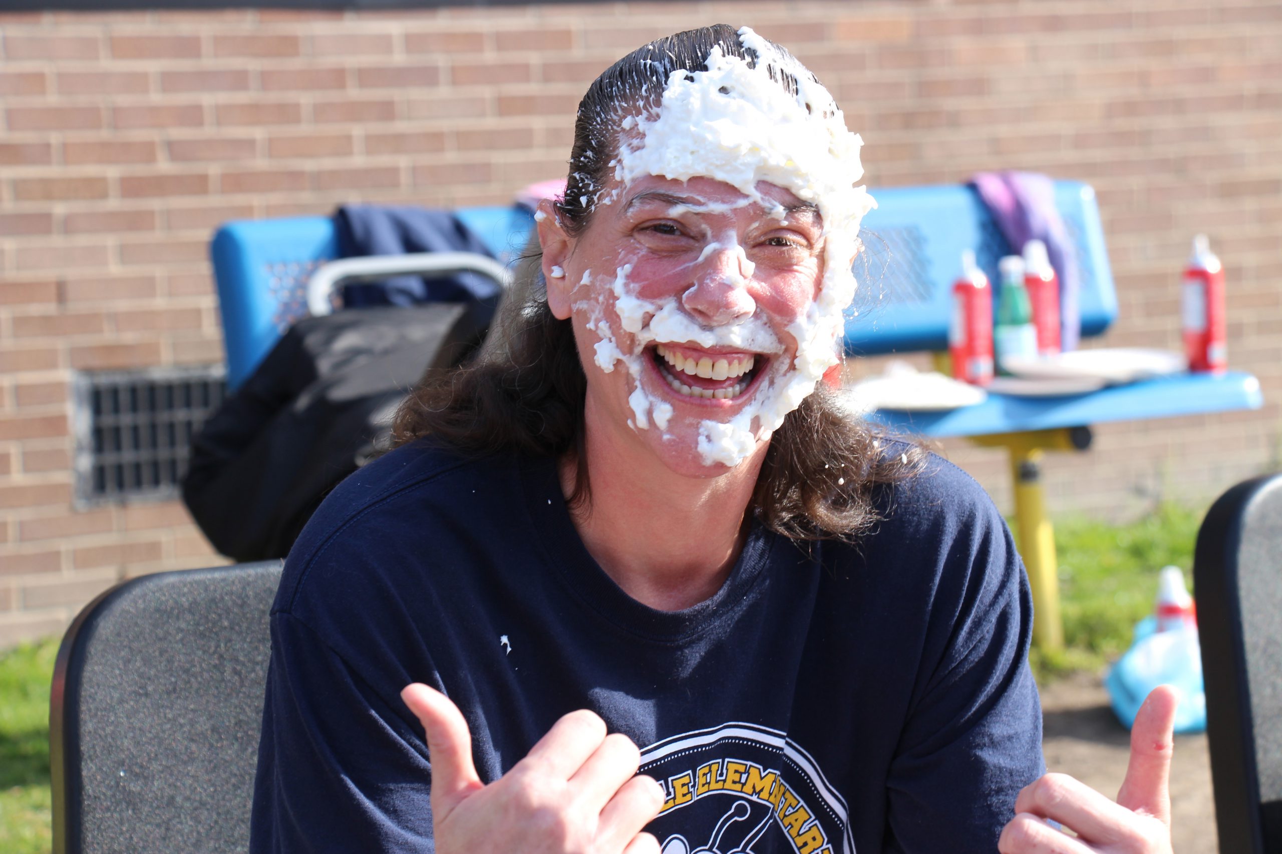 A woman in a navy blue shirt has whipped cream all over her face. She is smiling and giving a thumbs up.