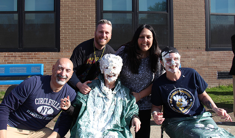 Two adults stand in back and three sit on chairs in the front. The three in front have whipped cream on their faces.